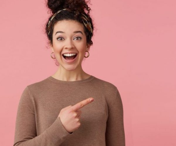 Portrait of happy amazed young beautiful lady with curly dark hair, heard the cool news, broadly smiling, looking at the camera, pointing with finger to copy space isolated over pink background.